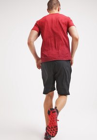 Camiseta deportiva roja con un patrón texturizado, combinada con pantalones cortos negros. Presenta zapatos negros y rojos con una suela flexible y una banda de rodadura detallada.