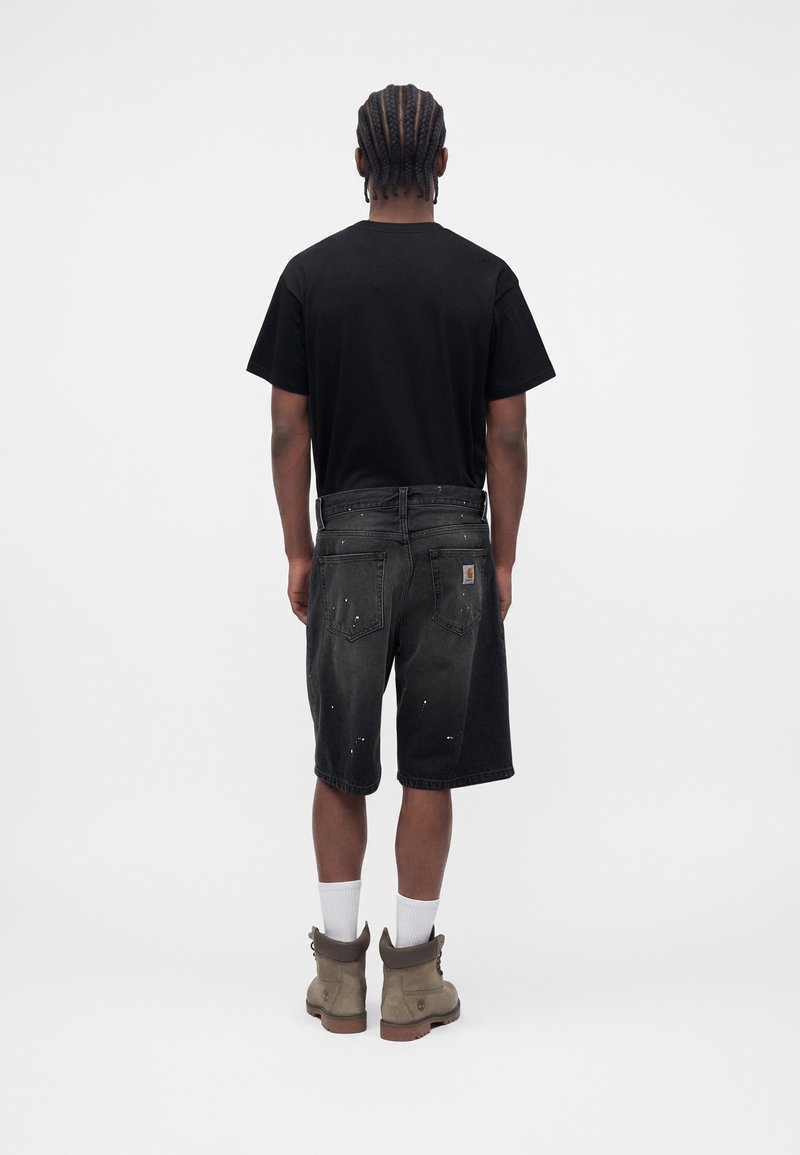 Man with braided hair wearing a black t-shirt, distressed black denim shorts, white socks, and beige work boots, standing facing away.
