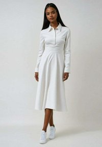 Robe blanche à manches longues avec col, présentant une taille cintrée et une jupe évasée. Tissu texturé avec des rayures subtiles. Associée à des baskets blanches.