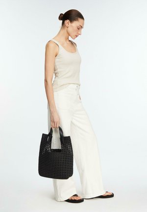 Woman in sleeveless beige top and white wide-leg pants holding a black woven tote bag, standing against a plain background.