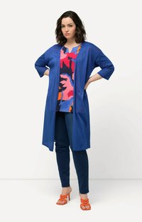 Modrý dlouhý cardigan s texturovanou tkaninou, nošený přes top s mnohobarevným abstraktním potiskem. Spárováno s tmavými skinny džíny a oranžovými sandály na podpatku.