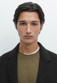 Uomo con capelli scuri e ondulati, che indossa un cappotto nero sopra un maglione verde oliva. Lo sfondo è bianco semplice. Il tono della pelle è chiaro.