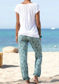 Persona de pie descalza en una playa de arena, vistiendo una camisa blanca de manga corta y pantalones con estampado floral en tonos azul-verde con los puños remangados.