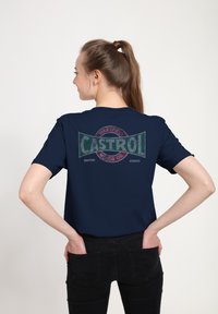 Námořnická modrá tričko s retro grafickým motivem Castrol v červené, zelené a bílé barvě na zadní straně, s krátkým rukávem a volným střihem.