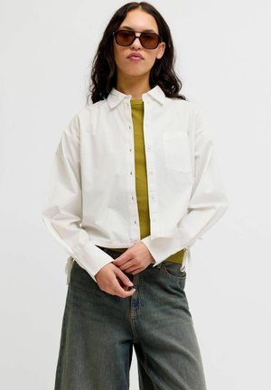 Žena s dlhými tmavými vlasmi, ktorá nosí tmavé slnečné okuliare, bielu košeľu s gombíkmi cez zelený top a voľné sivé džínsy, stojaca so zloženými rukami.