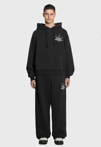 Czarna bluza z kapturem z przednią kieszenią i logo, zestawiona z luźnymi, czarnymi spodniami dresowymi z logo. Tkanina wydaje się miękka i teksturowana.
