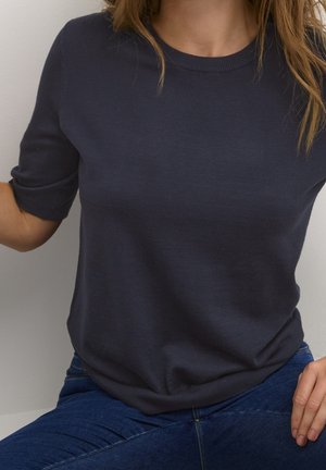 Kvinde iført en marineblå kortærmet striktop og blå jeans, siddende op ad en lysefarvet væg, med delvist synligt hår.