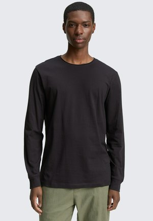 Joven con una camiseta negra de mangas largas y pantalones verde oliva, de pie contra un fondo liso gris claro.