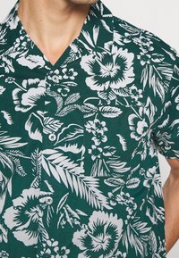 Lyhythihainen nappikauluspaita tummanvihreänä, jossa on valkoisia kukka- ja lehtikuvioita, kaulus ja tasku vasemmalla puolella.