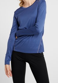 Camiseta deportiva de manga larga en azul, fabricada con tejido que absorbe la humedad. Presenta un cuello redondo y paneles laterales de malla para mayor transpirabilidad.