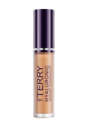 Anti-cernes By Terry Hyaluronic Serum dans un tube cylindrique transparent avec un bouchon violet foncé et un texte vertical sur le contenant.