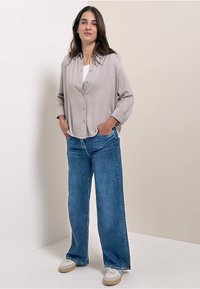 Chemise boutonnière gris clair à manches longues, assortie à un jean en denim bleu ample et des chaussures claires. Fond uni.