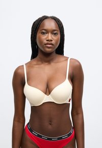 Béžová vystužená podprsenka s nastaviteľnými ramienkami a jemným logom. Zladená s červenými hipster nohavičkami s čiernym pásom s logom.