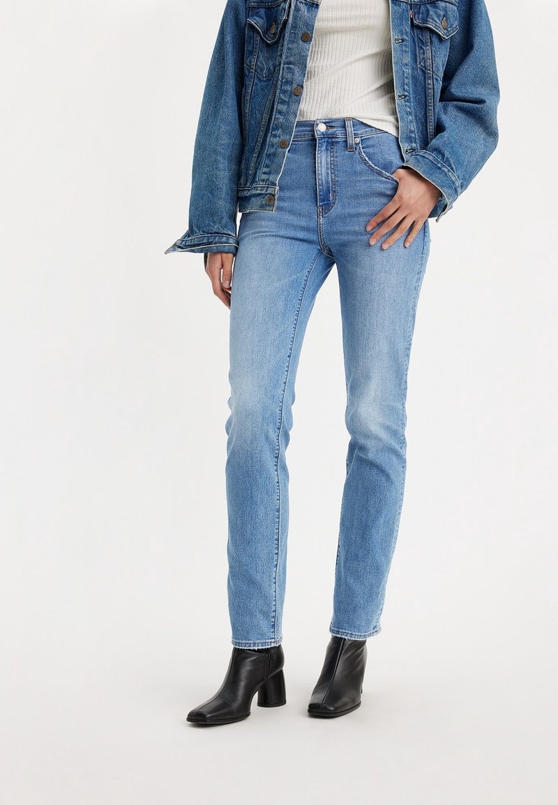 Femme portant un jean slim bleu clair, des bottines noires à talons, un haut côtelé blanc et une veste en jean bleue, debout avec une main près de la poche.