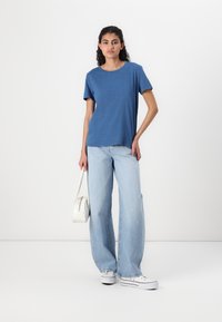 Blå t-skjorte med korte ermer og rund hals, sammen med lyseblå vide denimjeans. Modellen bærer en liten hvit håndveske.