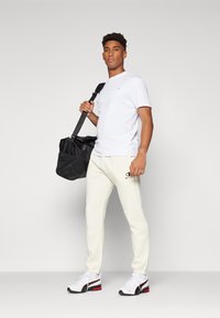 Chemise blanche à manches courtes, pantalon de survêtement crème avec logo, sac de sport noir et chaussures de sport blanches/rouges. Pose debout contre un fond gris.