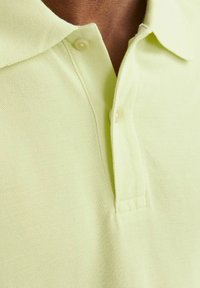 Svetle zelené polo tričko s límečkem, vyrobené z texturované tkaniny, se dvěma knoflíky a viditelným prošitím podél výstřihu.