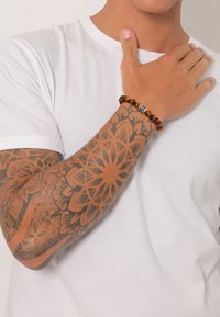 Pärlarmband med växlande mörkbruna och gyllene tigeröga-stenar, med en metallisk dödskalle som accent. Buren på en tatuerad underarm.