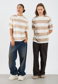 Two individuals stand side by side, wearing relaxed-fit t-shirts with beige and white stripes, paired with wide-leg jeans and casual footwear.