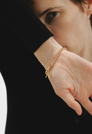 Persona que lleva una pulsera dorada con cuentas y un colgante en forma de "N", con la muñeca levantada cerca del rostro, vestido con una prenda de manga larga negra.