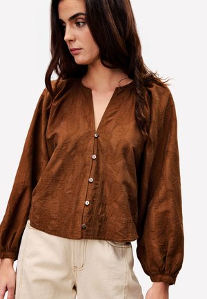 Femme aux longs cheveux bruns portant un chemisier marron à boutons avec des manches bouffantes et un pantalon taille haute beige clair.