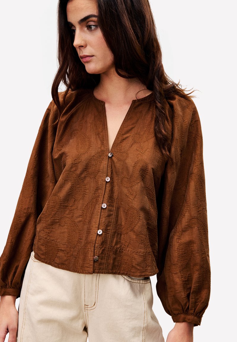 Frau mit langen braunen Haaren, die eine braune Bluse mit Knöpfen und Puffärmeln sowie hellbeige hochtaillierte Hose trägt.