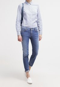 Kobieta w niebieskiej, striped koszuli na guziki, z podwiniętymi niebieskimi dżinsami, białych butach na luzie, niosąca ciemnoniebieski plecak na jednym ramieniu.