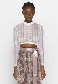 Weiße Spitzen-Crop-Top mit langen Ärmeln, kombiniert mit einem transparenten, hellrosa Rock mit Floralstickerei und einem Satinband zum Binden in der Taille.