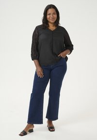 Black blouse with lace elbow sleeves, button-front design, paired with dark blue flared jeans and black block-heeled sandals.