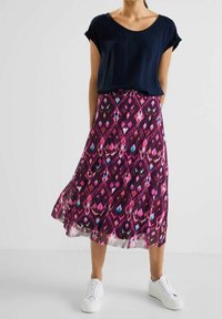 Kvinna iklädd en flödande midi-kjol i lila och rosa mönster, marinblå kortärmad topp och vita sneakers som står på ett vitt golv.