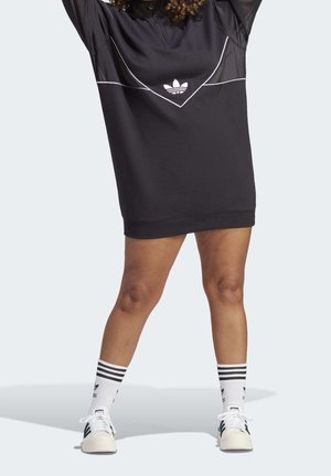 Osoba ubrana w czarną sukienkę Adidas z białym logo, białe skarpety Adidas z czarnymi paskami oraz białe buty sportowe Adidas, stojąca z nogami rozstawionymi.