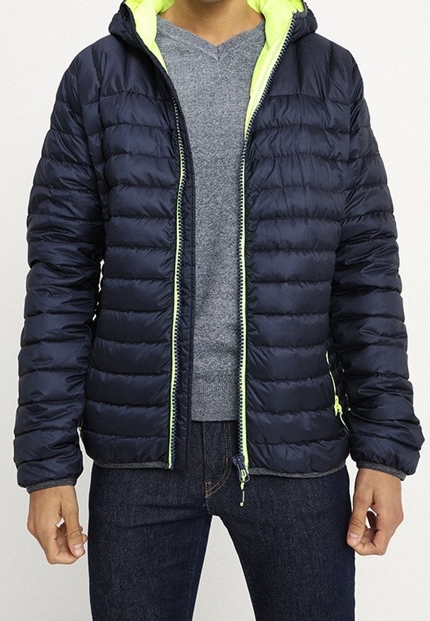 Personne portant une veste matelassée bleu marine avec une doublure vert néon, un pull gris et un jean foncé, se tenant avec les mains détendues sur les côtés.