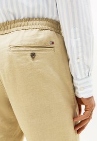 Beige Leinenhosen mit elastischem Bund, einer geknöpften Gesäßtasche und einem kleinen rot-weiß-blauen Logo, getragen mit einem hellblauen gestreiften Hemd.