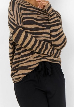 Persona che indossa un maglione a righe zebra beige e nere con pantaloni neri, mano posata pensierosamente vicino al viso, su sfondo bianco.