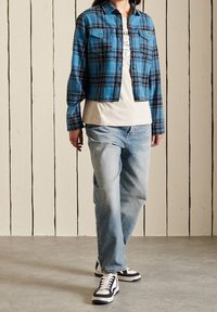 Camicia di flanella a quadri blu cropped con due tasche sul petto, indossata sopra una maglietta chiara e abbinata a jeans azzurri chiari e sneakers nere.