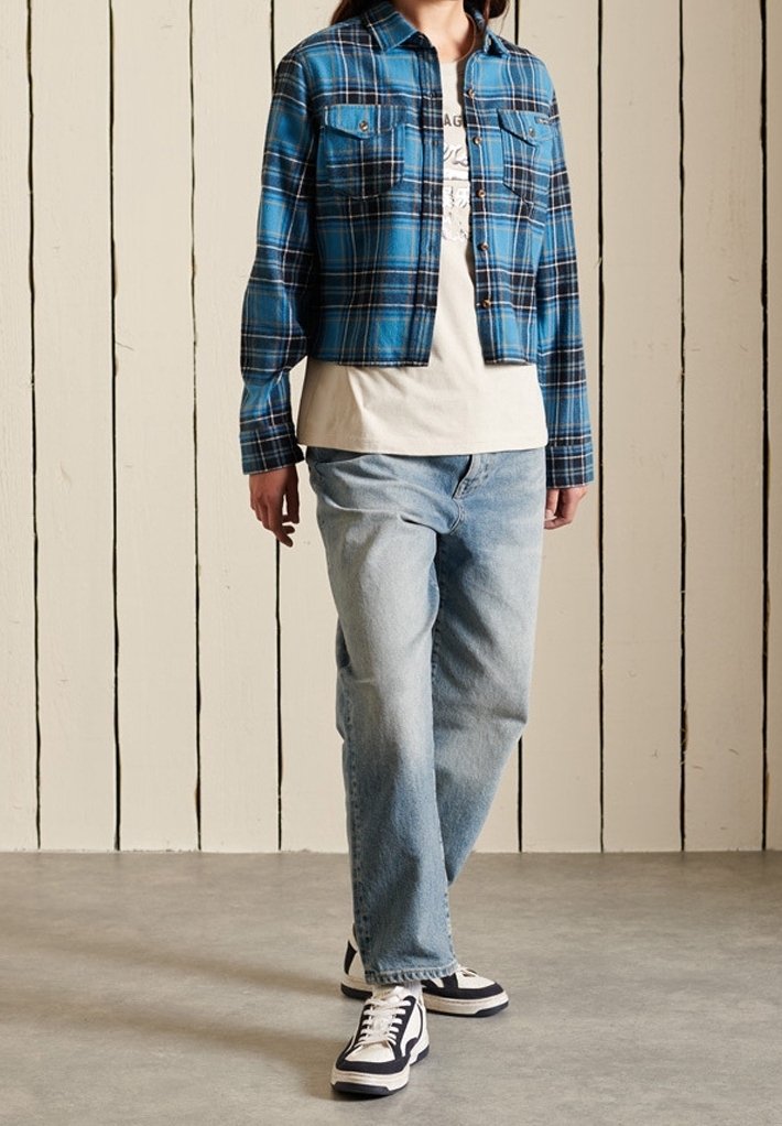 Camicia di flanella a quadri blu cropped con due tasche sul petto, indossata sopra una maglietta chiara e abbinata a jeans azzurri chiari e sneakers nere.