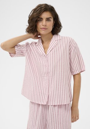 Młoda kobieta o falowanych brązowych włosach, ubrana w różowo-białą, pionowo prążkowaną koszulę z krótkim rękawem i pasujące spodnie, pozująca z ręką przy szyi.