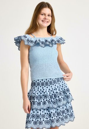 Nuori nainen esittelee vaaleansinisen, frillatun yläosan ja siihen sopivan kerroksellisen hameen, joissa on tumma kukkakirjonta, hymyillen yksiväristä taustaa vasten.