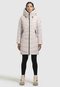 Padded coat in light beige with a high collar, front zipper, and angled quilting. Paired with black leggings and beige boots.
