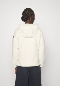 Persona con capelli intrecciati che indossa un felpa di pile bianca con testo ricamato sul cappuccio e pantaloni neri, di spalle rispetto alla telecamera.