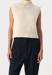 Chaleco de punto color crema con cuello alto, textura acanalada, mangas cortas y un dobladillo corto, combinado con pantalones azul marino.