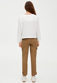 Blouse blanche à manches longues, légèrement transparente, avec un col arrondi ; associée à un pantalon cargo marron avec poches latérales et chevilles.