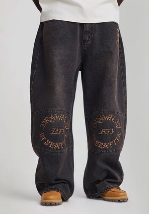 Jeans noirs baggy avec texte brodé beige "Drawn by ED in Seattle" sur les genouillères, portés avec des bottes beiges et une chemise blanche.