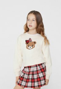 Felpa crema con una faccia di orso e un fiocco scozzese, abbinata a una gonna plissettata scozzese rossa. Tessuto morbido e vestibilità comoda.