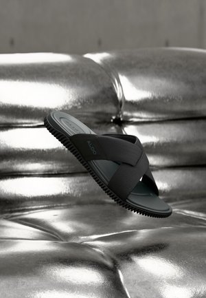 Svart slidesandal med kryssede stropper som flyter over en skinnende, metallisk sølv polstret overflate.