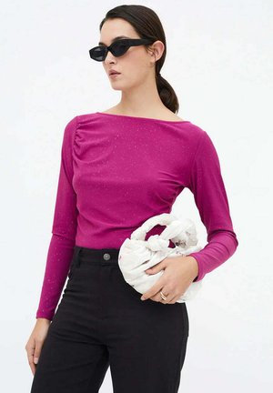 Mujer con gafas de sol negras, blusa de manga larga magenta, pantalones negros, sosteniendo un pequeño bolso blanco anudado a la altura de la cintura.