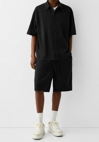 Osoba v volné černé košili s krátkým rukávem na knoflíky a odpovídajících černých šortkách, doplněná bílými ponožkami a bílými teniskami.