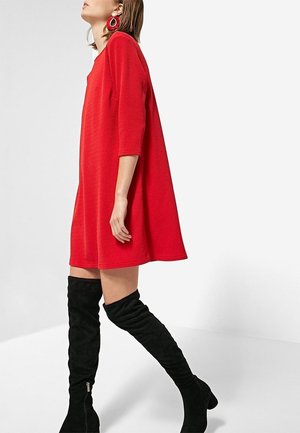 Femme en robe rouge ample à manches trois-quarts, portant des bottes noires au-dessus du genou, debout devant un fond blanc uni.