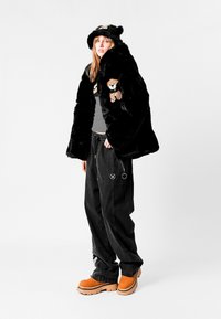 Chaqueta de piel sintética negra con parches de oso de peluche, combinada con pantalones negros holgados y botas de plataforma naranjas. Los accesorios incluyen un sombrero de oso negro.