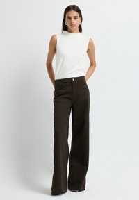 Haut sans manches blanc associé à un pantalon évasé marron foncé orné de petites embellissements. Les deux vêtements ont une texture lisse et une coupe ajustée.